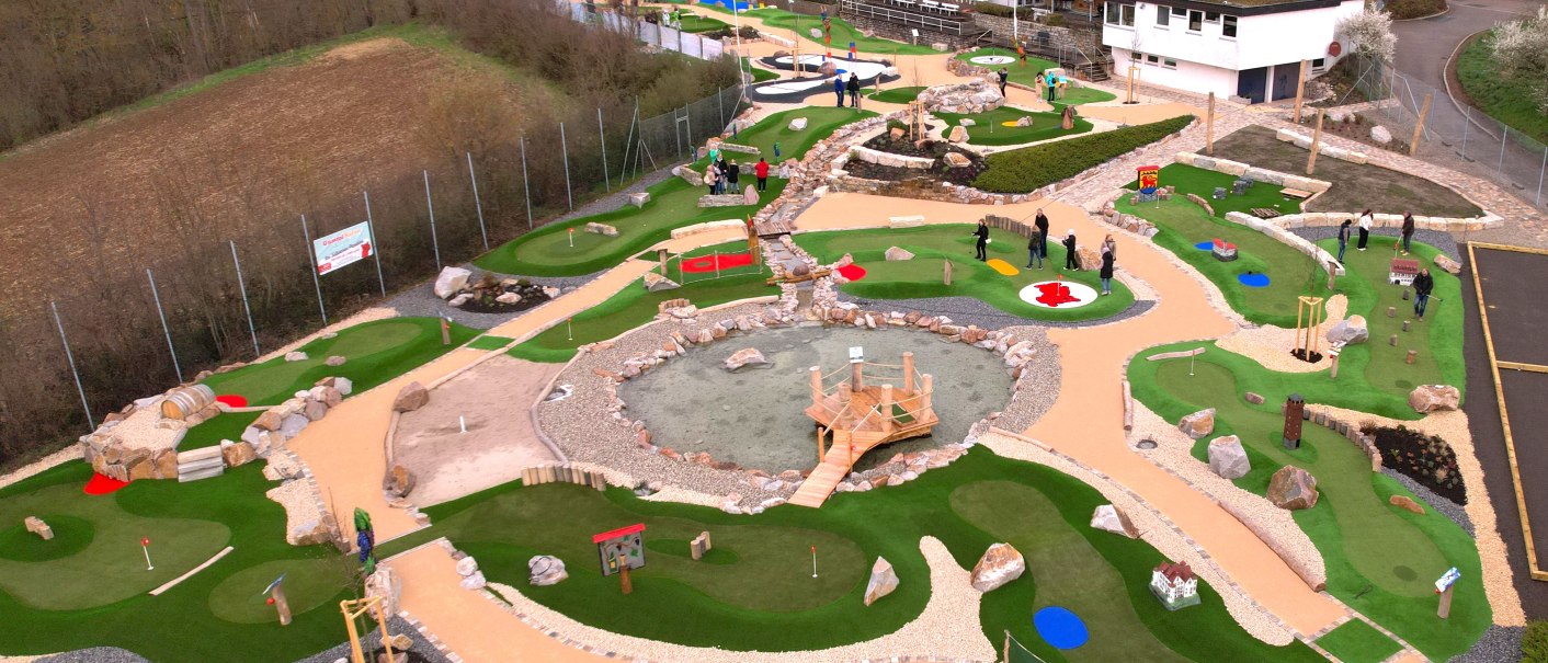 Luftaufnahme eines Adventure-Golfplatzes in Vaihingen mit grünen Bahnen, Hindernissen und einem zentralen Teich. Menschen spielen auf dem Platz., © TV Vaihingen/Enz