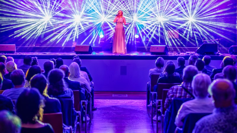 Eine S&auml;ngerin steht auf der B&uuml;hne der Stadthalle K3N, umgeben von bunten Lichteffekten. Das Publikum sitzt in Reihen und schaut zu., &copy; Daniel Jueptner | 8pm.de