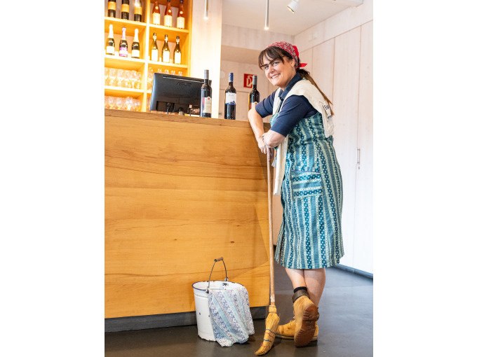 Frau in traditioneller Kleidung lehnt lächelnd an einer Theke mit Weinflaschen. Ein Eimer und Besen stehen daneben., © Stuttgart Marketing GmbH Frau in traditioneller Kleidung lehnt lächelnd an einer Theke mit Weinflaschen. Ein Eimer und Besen stehen daneben., © Stuttgart Marketing GmbH