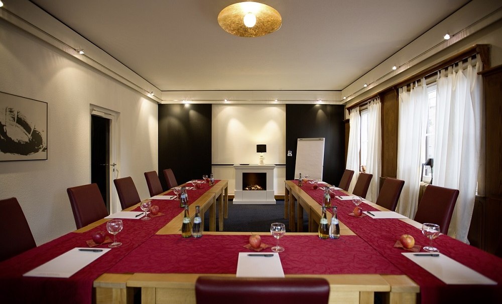 Konferenzraum mit U-f&ouml;rmigem Tisch, roten St&uuml;hlen, Notizbl&ouml;cken, Wasserflaschen und &Auml;pfeln. Ein Kamin und Flipchart im Hintergrund., &copy; Hotel B&uuml;rkle