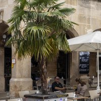 Caf&eacute; mit Tischen und St&uuml;hlen unter Sonnenschirmen vor einem historischen Geb&auml;ude mit Arkaden. Palmen und Menschen im Au&szlig;enbereich., &copy; SMG, Sarah Schmid