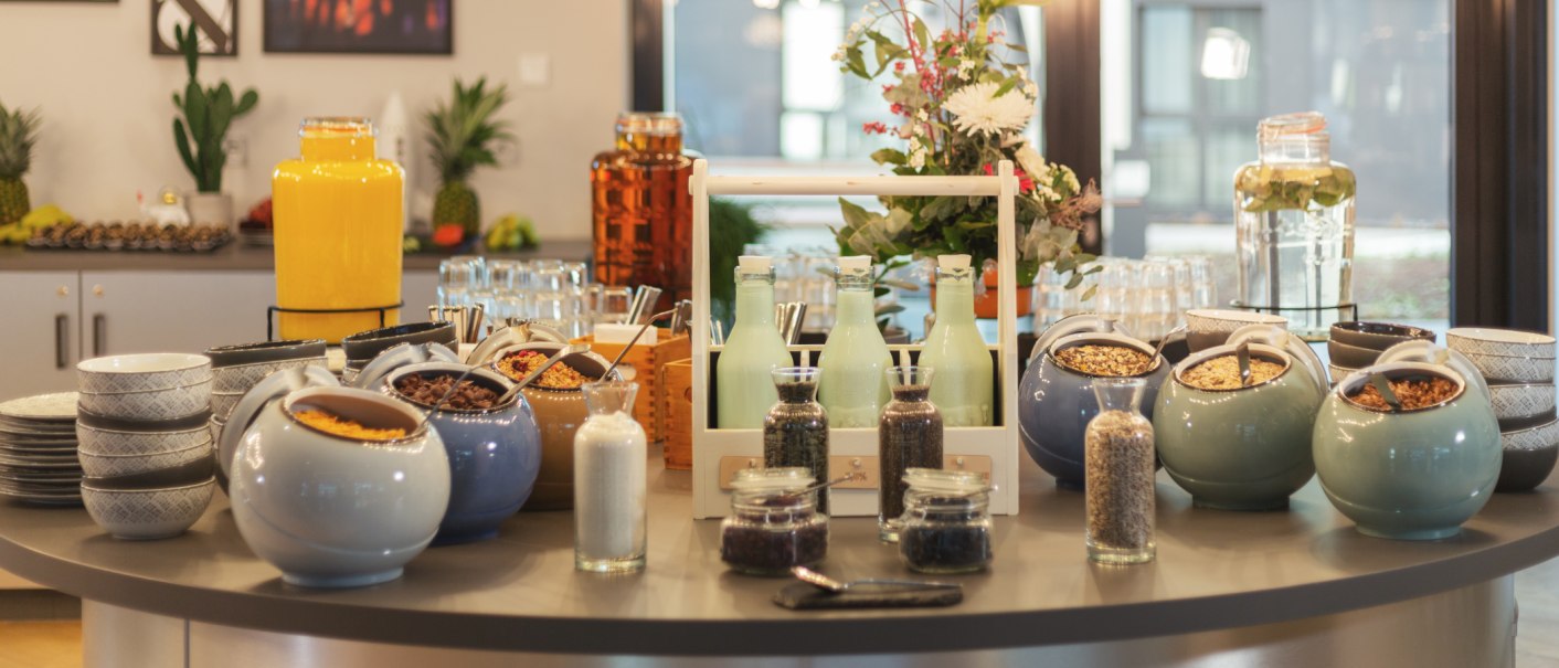 Ein Frühstücksbuffet mit verschiedenen Müslisorten, Milchflaschen und Saftspendern. Im Hintergrund sind Blumen und Gläser zu sehen., © Novum Management GmbH Ein Frühstücksbuffet mit verschiedenen Müslisorten, Milchflaschen und Saftspendern. Im Hintergrund sind Blumen und Gläser zu sehen., © Novum Management GmbH