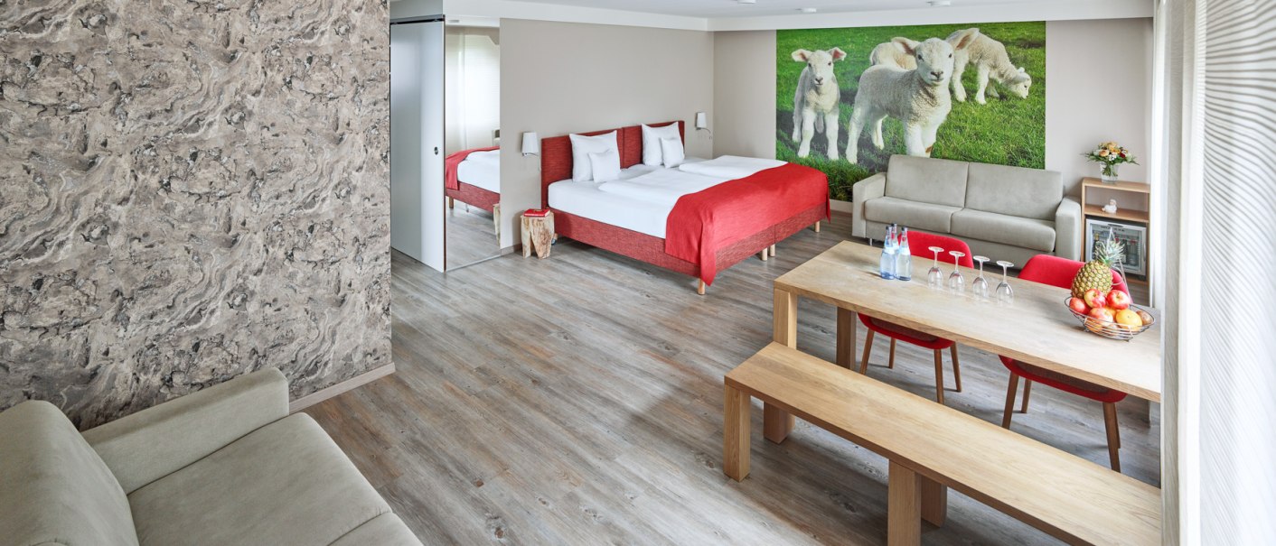 Modernes Zimmer mit Doppelbett, roter Bettdecke, Sofa, Esstisch und Wandbild von Lämmern. Helle, freundliche Einrichtung mit Holzfußboden., © Hotel Restaurant Lamm