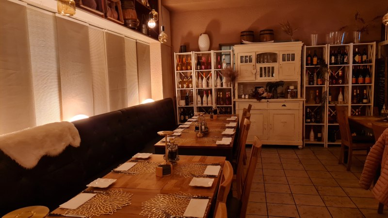 Ein gem&uuml;tlicher Restaurantinnenraum mit gedeckten Holztischen, dekorativen Platzsets und warmem Licht. Regale mit Flaschen und ein rustikaler Schrank erg&auml;nzen das Ambiente., &copy; RistoRocco