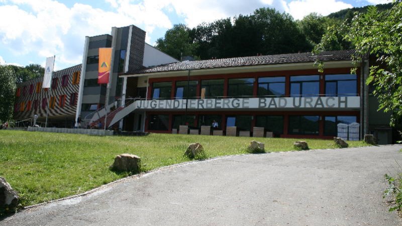 Die Jugendherberge Bad Urach mit moderner Architektur, bunten Fenstern und einem großen Schriftzug. Vorne ein gepflegter Rasen und ein Weg. Die Jugendherberge Bad Urach mit moderner Architektur, bunten Fenstern und einem großen Schriftzug. Vorne ein gepflegter Rasen und ein Weg.