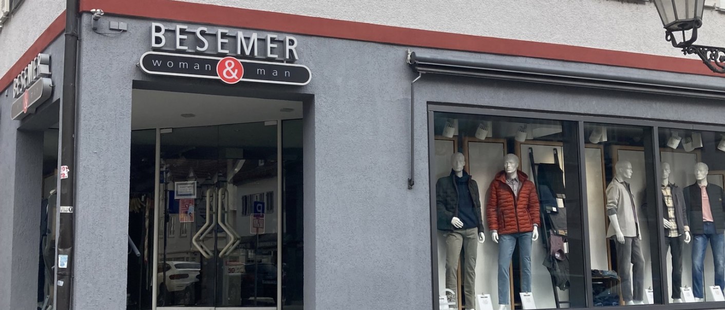 Modehaus Besemer in Nürtingen mit Schaufensterpuppen und Eingangstür. Graue Fassade mit roten Akzenten, Kopfsteinpflaster davor., © Lena Besemer Modehaus Besemer in Nürtingen mit Schaufensterpuppen und Eingangstür. Graue Fassade mit roten Akzenten, Kopfsteinpflaster davor., © Lena Besemer
