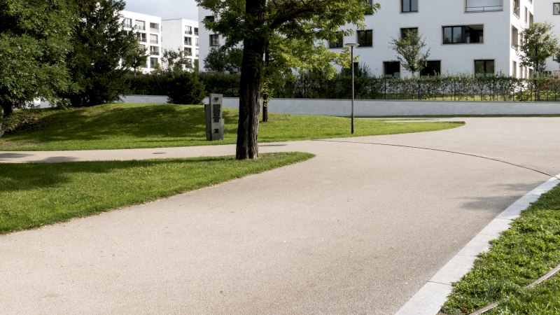 Ein gepflegter Park mit geschwungenen Gehwegen, Bäumen und modernen Wohngebäuden im Hintergrund. Ein Gedenkstein ist sichtbar., © Foto-Kunst Andreas Keller Ein gepflegter Park mit geschwungenen Gehwegen, Bäumen und modernen Wohngebäuden im Hintergrund. Ein Gedenkstein ist sichtbar., © Foto-Kunst Andreas Keller