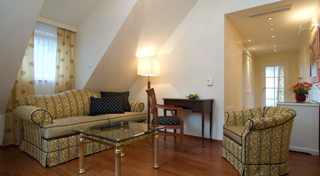 Eleganter Wohnbereich mit gemustertem Sofa, Sessel, Glastisch und Schreibtisch. Warme Beleuchtung und Holzboden schaffen eine gemütliche Atmosphäre., © Jeanette Bender Eleganter Wohnbereich mit gemustertem Sofa, Sessel, Glastisch und Schreibtisch. Warme Beleuchtung und Holzboden schaffen eine gemütliche Atmosphäre., © Jeanette Bender