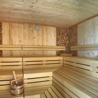 Innenansicht einer Sauna aus hellem Holz mit Bänken, einem Holzeimer und gestapeltem Holz an den Wänden., © TOMAS