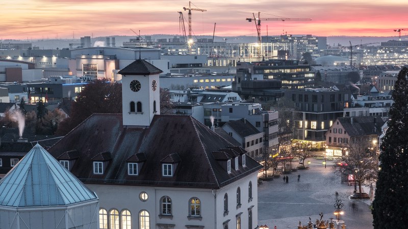 Stadtansicht bei Sonnenuntergang: Ein Kirchturm im Vordergrund, moderne Gebäude und Baukräne im Hintergrund, beleuchtete Fenster und ein farbenfroher Himmel., © Stadt Sindelfingen
