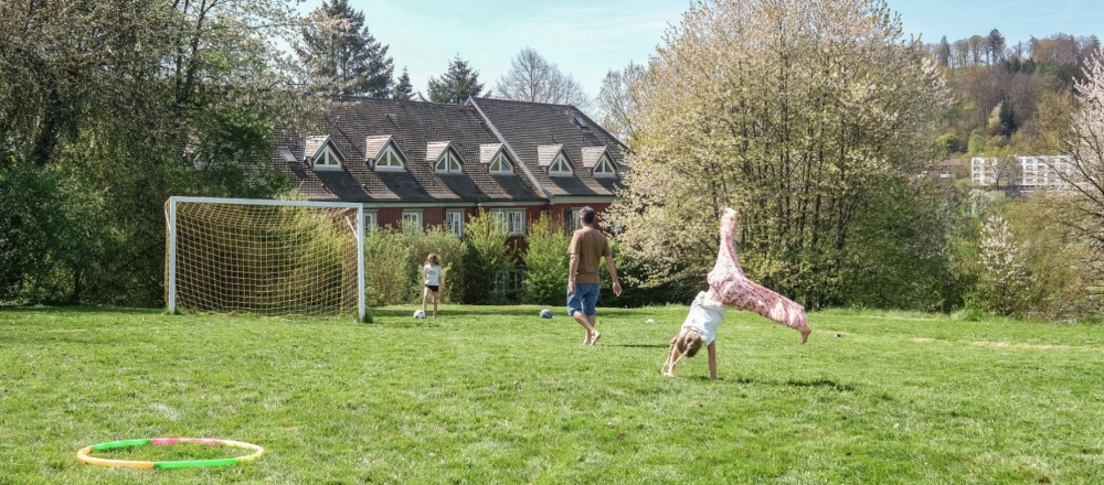 Kinder spielen auf einem grünen Fußballplatz mit einem Tor, umgeben von Bäumen und einem Haus im Hintergrund., © DJH Baden-Württemberg e.V.