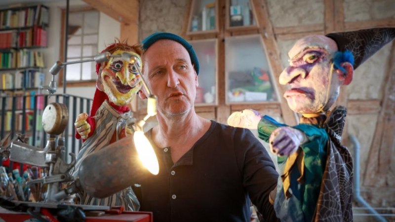 Ein Mann mit zwei Handpuppen, darunter Kasperl, in einem Raum mit Bücherregalen. Die Puppen sind detailreich gestaltet., © Bildrechte: Gregor Oehmann Ein Mann mit zwei Handpuppen, darunter Kasperl, in einem Raum mit Bücherregalen. Die Puppen sind detailreich gestaltet., © Bildrechte: Gregor Oehmann