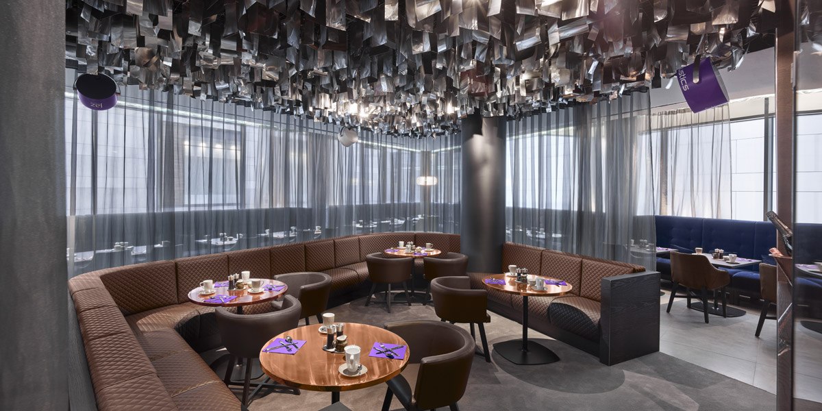 Modernes Restaurant mit runden Holztischen, braunen Stühlen und dekorativer Decke. Gedämpftes Licht und Vorhänge schaffen eine gemütliche Atmosphäre., © Jaz Hotel Stuttgart
