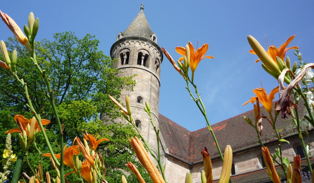 Turm des Klosters Lorch ragt in den blauen Himmel, umgeben von grünen Bäumen und orangefarbenen Lilien im Vordergrund., © Remstal Tourismus e.V.