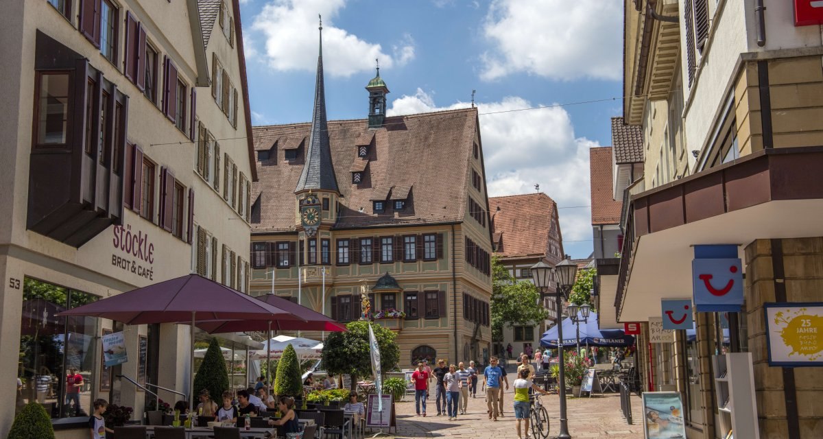 Belebte Fu&szlig;g&auml;ngerzone mit historischen Geb&auml;uden, Caf&eacute; und Menschen unter Sonnenschirmen. Im Hintergrund ein Geb&auml;ude mit Turm und Uhr., &copy; Stuttgart Marketing GmbH Achim Mende