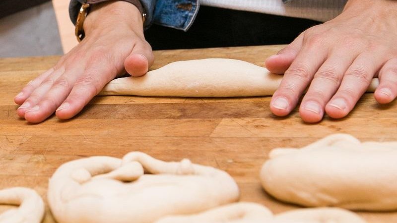Eine Person formt Teig auf einem Holzbrett. Mehrere Teigstücke liegen bereit. Die Person trägt eine Jeansjacke., © © T&E, Benjamin Stollenberg Eine Person formt Teig auf einem Holzbrett. Mehrere Teigstücke liegen bereit. Die Person trägt eine Jeansjacke., © © T&E, Benjamin Stollenberg
