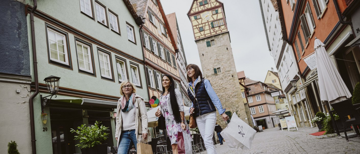 Drei Frauen schlendern mit Einkaufstaschen durch eine malerische Straße in Schwäbisch Hall, umgeben von Fachwerkhäusern und einem Turm., © Nico Kurth