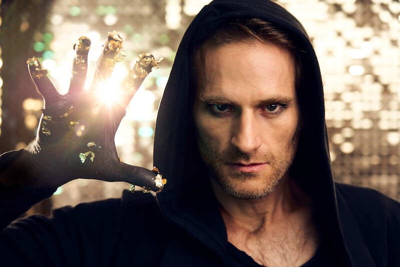 Ein Mann in einem schwarzen Kapuzenpullover h&auml;lt seine Hand mit goldenen Akzenten in die Kamera. Licht scheint durch seine Finger und erzeugt einen magischen Effekt., &copy; EMT Event-Media-Tec GmbH