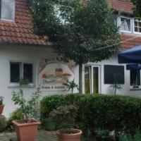 Ein gemütliches Restaurant mit roten Dachziegeln, umgeben von Pflanzen. Ein blauer Sonnenschirm steht im Vordergrund.