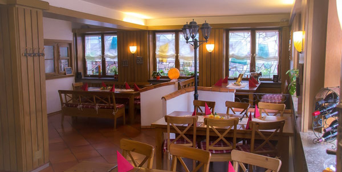 Gemütliche Gaststube mit Holzmöbeln, dekorierten Tischen und roten Servietten. Große Fenster und warme Beleuchtung schaffen eine einladende Atmosphäre.