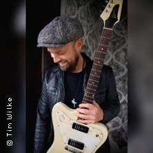 Ein Mann mit Mütze und Lederjacke hält eine E-Gitarre und lächelt nach unten., © links im Bild Ein Mann mit Mütze und Lederjacke hält eine E-Gitarre und lächelt nach unten., © links im Bild
