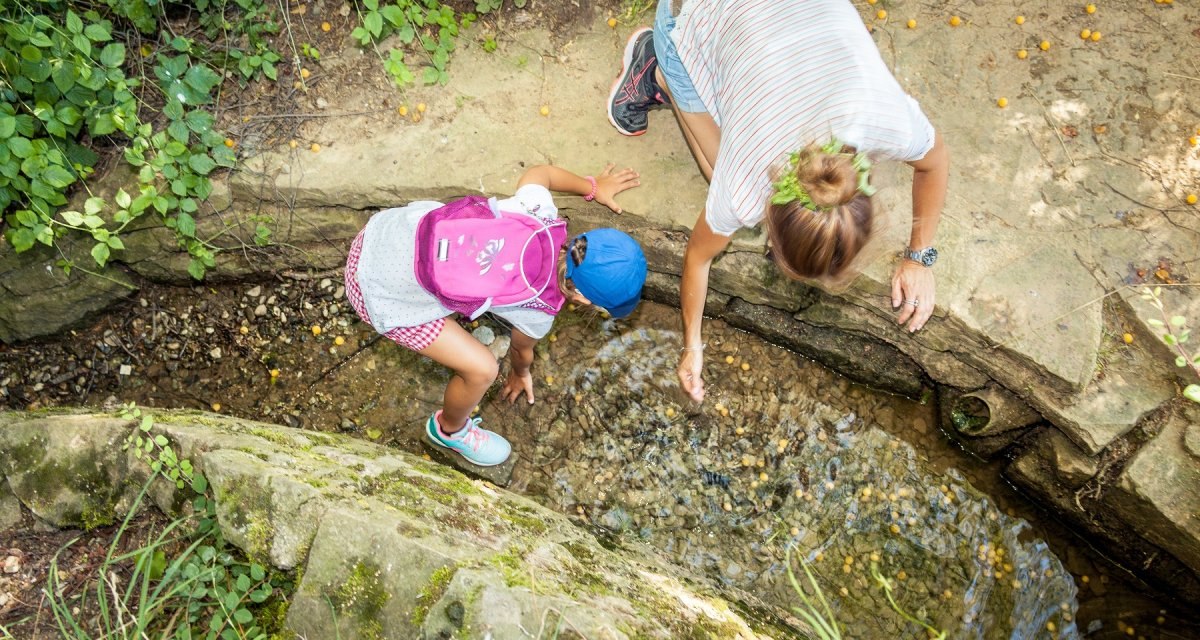 Ein Kind mit rosa Rucksack und ein Erwachsener beugen sich über einen kleinen Bach im Freien, um im Wasser zu spielen., © hochgehberge Ein Kind mit rosa Rucksack und ein Erwachsener beugen sich über einen kleinen Bach im Freien, um im Wasser zu spielen., © hochgehberge
