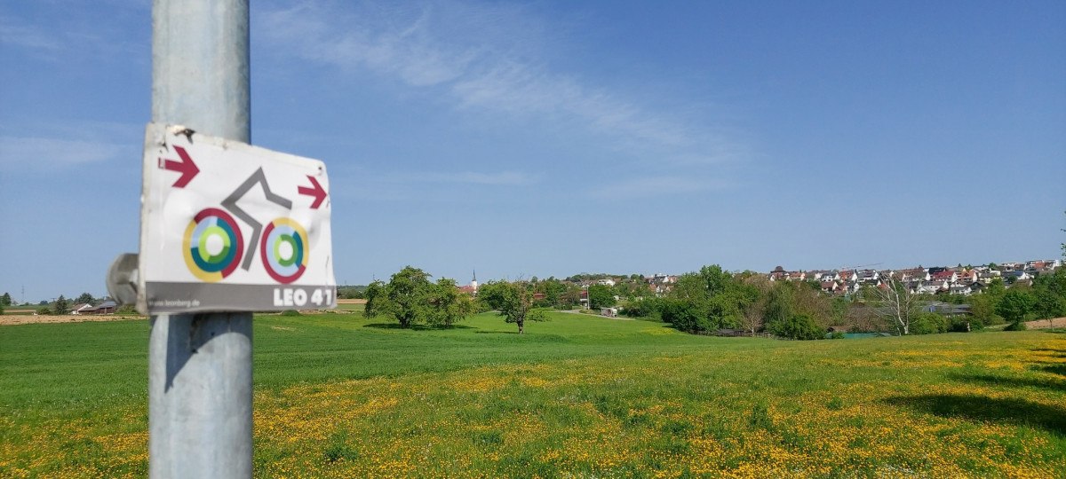Ein Fahrradweg-Schild an einem Pfosten vor einer gr&uuml;nen Wiese mit gelben Blumen. Im Hintergrund ist ein Dorf zu sehen., &copy; Natur.Nah. Sch&ouml;nbuch & Heckeng&auml;u
