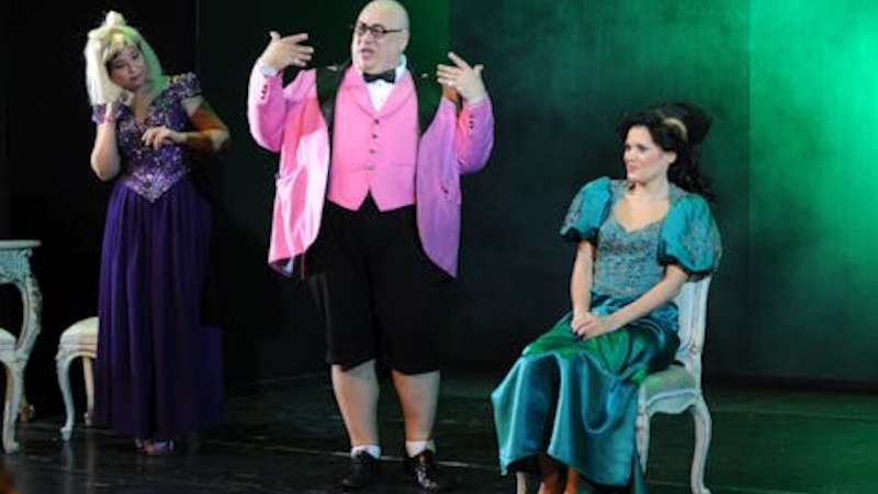 Drei Darsteller in farbenfrohen Kost&uuml;men auf einer B&uuml;hne. Eine Person steht gestikulierend, w&auml;hrend die anderen beiden sitzen und zuschauen., &copy; Touristik Bad Wildbad GmbH