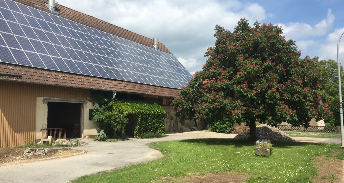 Ein Bauernhof mit Solarpanelen auf dem Dach. Ein blühender Baum steht im Vordergrund auf einer grünen Wiese unter blauem Himmel., © www.pro-cycl.de Ein Bauernhof mit Solarpanelen auf dem Dach. Ein blühender Baum steht im Vordergrund auf einer grünen Wiese unter blauem Himmel., © www.pro-cycl.de