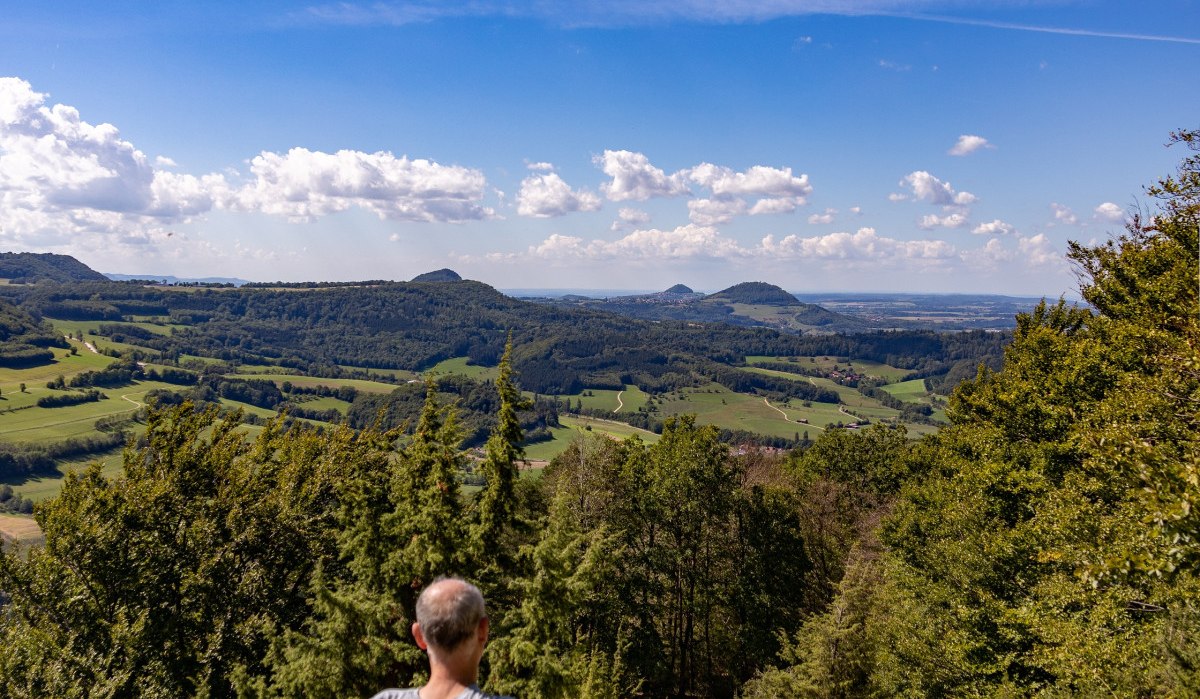 Eine Person sitzt auf einer Bank und blickt auf eine weite, grüne Landschaft unter einem klaren, blauen Himmel mit wenigen Wolken., © Foto Thomas Zehnder Eine Person sitzt auf einer Bank und blickt auf eine weite, grüne Landschaft unter einem klaren, blauen Himmel mit wenigen Wolken., © Foto Thomas Zehnder