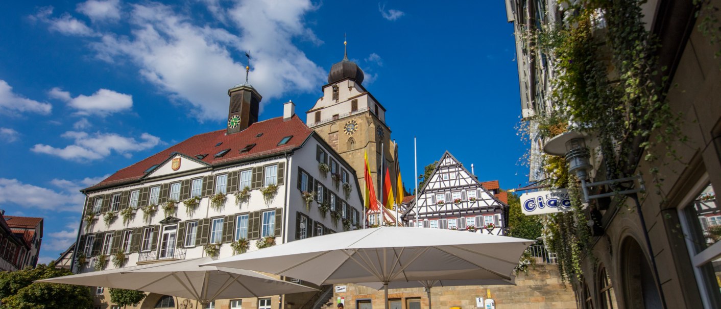 Historische Gebäude in Herrenberg mit Fachwerkhaus, Kirchturm und Sonnenschirmen. Blauer Himmel und bunte Fahnen im Hintergrund., © SMG Achim Mende Historische Gebäude in Herrenberg mit Fachwerkhaus, Kirchturm und Sonnenschirmen. Blauer Himmel und bunte Fahnen im Hintergrund., © SMG Achim Mende