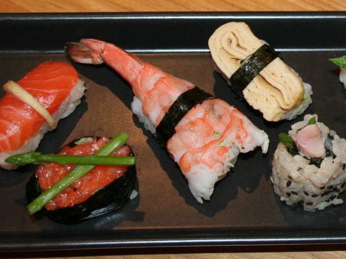 Ein schwarzer Teller mit verschiedenen Sushi-Varianten, darunter Lachs, Garnele, Omelett und Reisrollen, dekoriert mit Spargel und Algen., &copy; Kultweine-Kochwelten-Kunst&uuml;cke - Kochschule Sonja Lenz