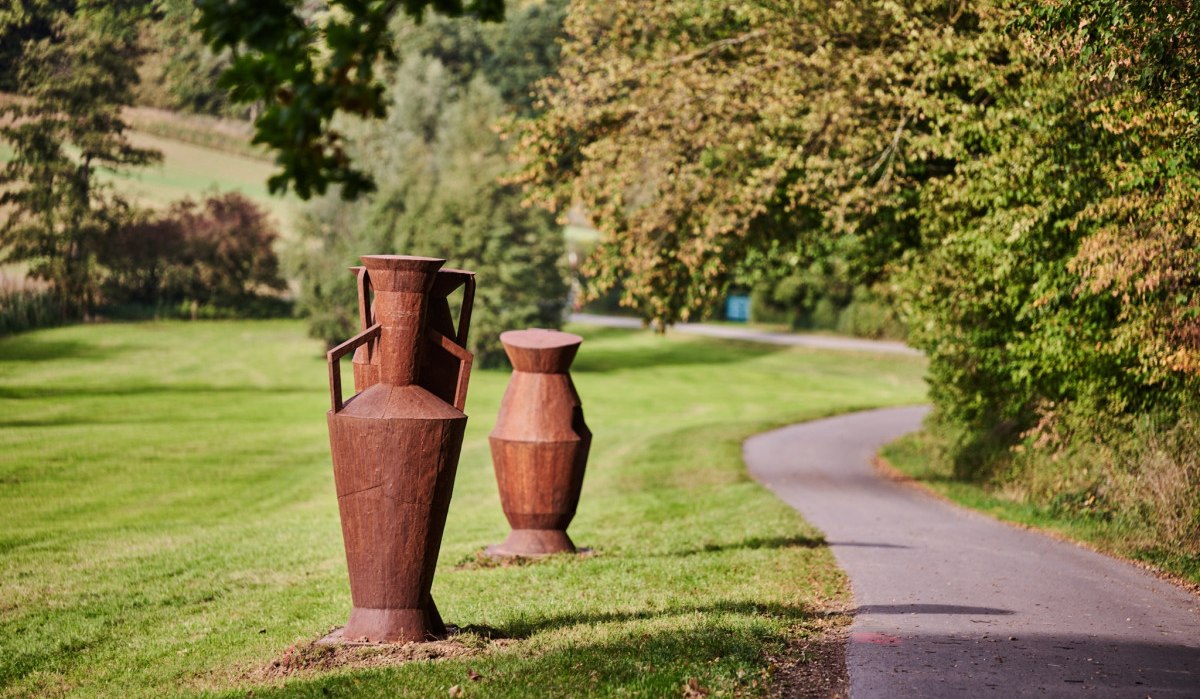 Zwei rostfarbene Amphoren-Skulpturen auf einer Wiese neben einem Weg, umgeben von Bäumen., © Natur.Nah. Schönbuch & Heckengäu Zwei rostfarbene Amphoren-Skulpturen auf einer Wiese neben einem Weg, umgeben von Bäumen., © Natur.Nah. Schönbuch & Heckengäu