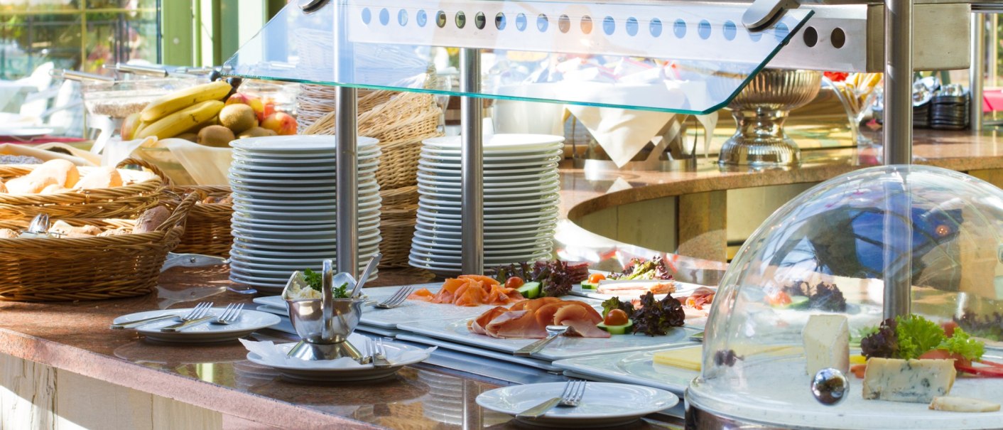 Ein reichhaltiges Buffet mit Tellern, Obst, Brot und Käse unter einer Glashaube in einem hellen Raum mit Pflanzen im Hintergrund., © TOMAS