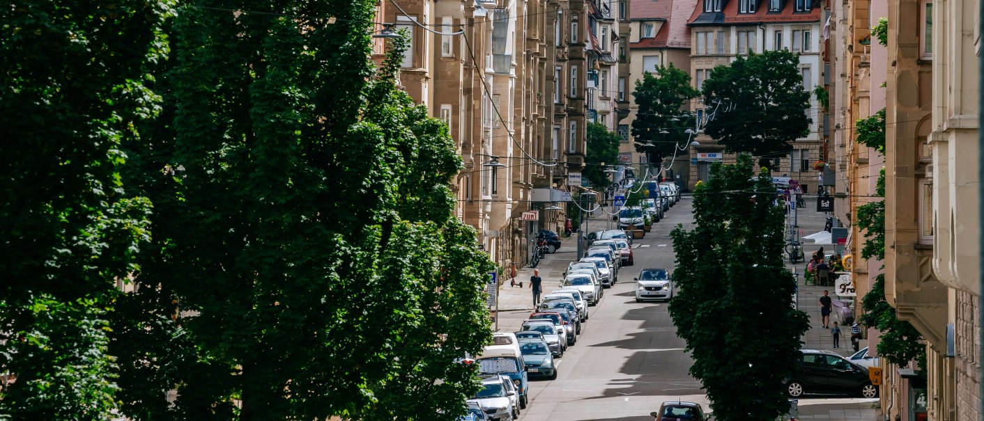 Eine belebte Stadtstraße mit geparkten Autos, alten Gebäuden und Bäumen. Im Hintergrund sind Hügel und weitere Häuser zu sehen., © Thomas Niedermüller
