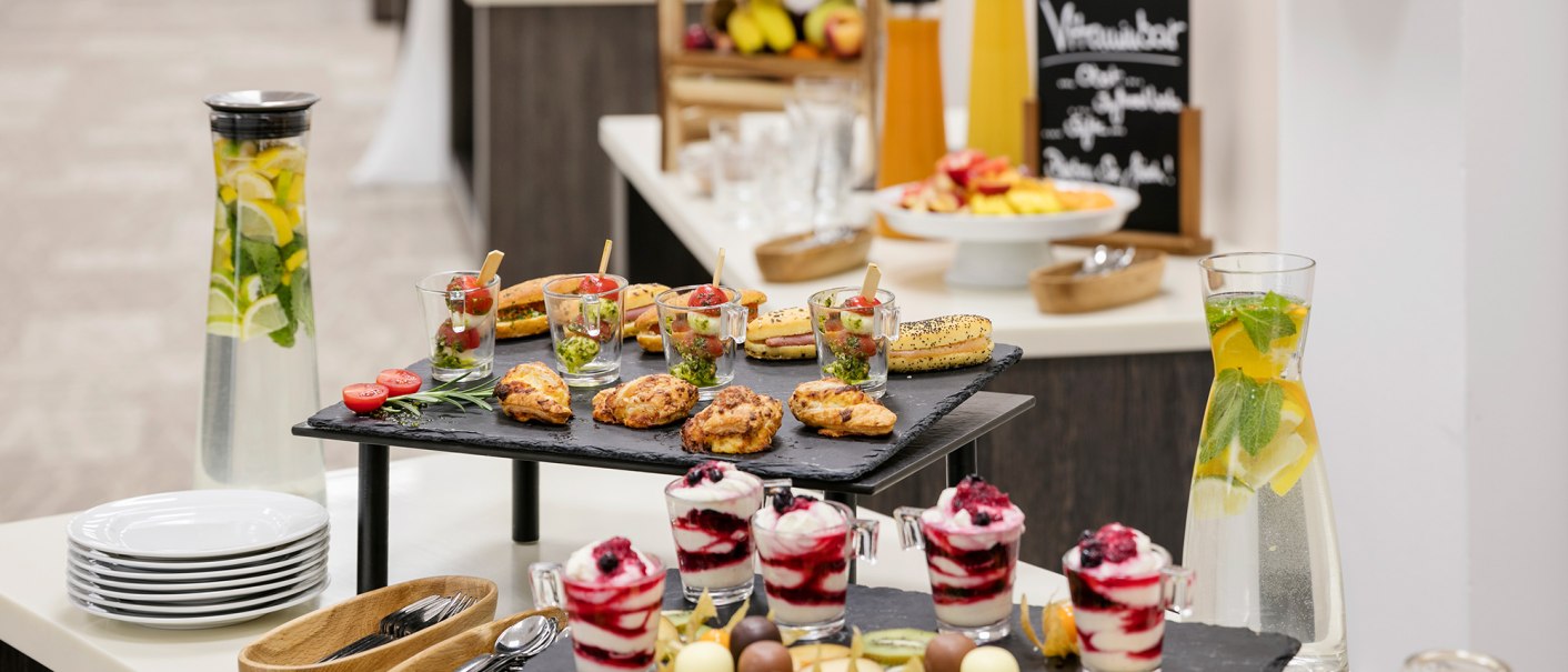 Ein Buffet mit verschiedenen Snacks, Desserts und Getr&auml;nken. Im Vordergrund stehen Gl&auml;ser mit Wasser und Zitronenscheiben, daneben Teller und Besteck., &copy; ARCOTEL Hotels