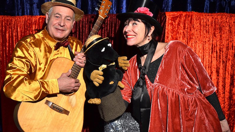 Zwei Personen in bunten Kostümen posieren mit einer Maulwurf-Puppe und Gitarre vor einem roten Vorhang., © FILharmonie Filderstadt