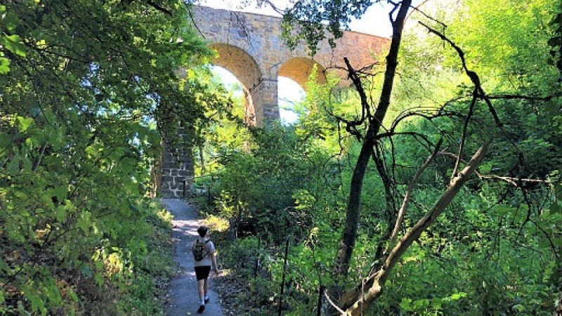 Ein Wanderer auf einem schattigen Pfad unter einer steinernen Bogenbrücke, umgeben von dichtem Grün., © Touristik und Marketing GmbH Ein Wanderer auf einem schattigen Pfad unter einer steinernen Bogenbrücke, umgeben von dichtem Grün., © Touristik und Marketing GmbH