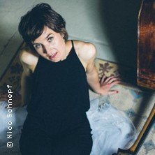 Eine Frau in schwarzem Outfit sitzt auf einem Teppich neben einem Kontrabass, schaut nach oben. Der Raum ist dezent beleuchtet., &copy; links im Bild