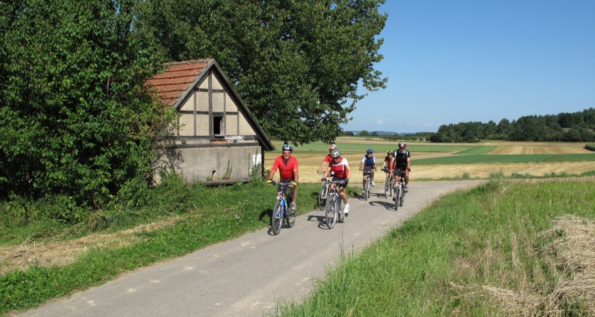 Vier Radfahrer fahren auf einem ländlichen Weg an einem kleinen Fachwerkhaus vorbei. Die Landschaft ist grün und weitläufig, der Himmel klar und blau., © Natur.Nah. Schönbuch & Heckengäu Vier Radfahrer fahren auf einem ländlichen Weg an einem kleinen Fachwerkhaus vorbei. Die Landschaft ist grün und weitläufig, der Himmel klar und blau., © Natur.Nah. Schönbuch & Heckengäu