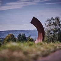 Gro&szlig;e, gebogene Skulptur in einer Landschaft mit B&auml;umen und H&uuml;geln im Hintergrund unter blauem Himmel., &copy; Natur.Nah. Sch&ouml;nbuch & Heckeng&auml;u