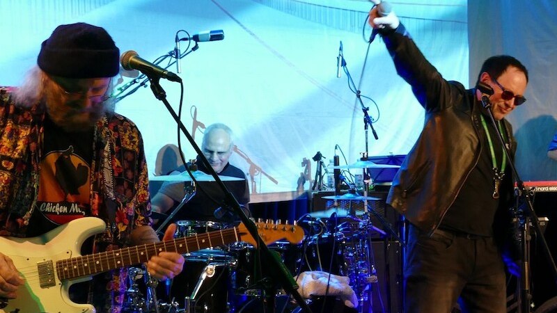 Eine Band performt auf einer Bühne. Ein Gitarrist spielt E-Gitarre, ein Schlagzeuger sitzt am Schlagzeug, und ein Sänger mit Sonnenbrille singt leidenschaftlich., © Mauerwerk GmbH
