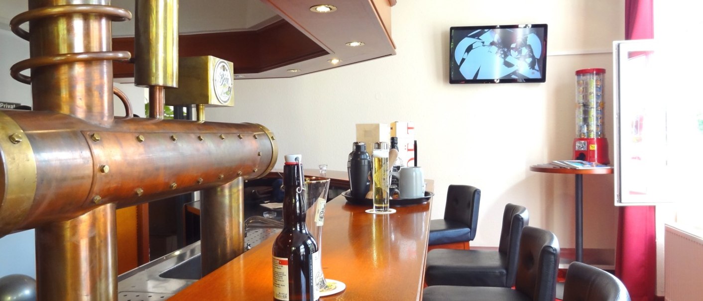 Eine Bar mit hölzerner Theke, Barhockern und einem Fernseher an der Wand. Ein auffälliger Zapfhahn aus Metall ist zu sehen., © Hotel Arcis Eine Bar mit hölzerner Theke, Barhockern und einem Fernseher an der Wand. Ein auffälliger Zapfhahn aus Metall ist zu sehen., © Hotel Arcis