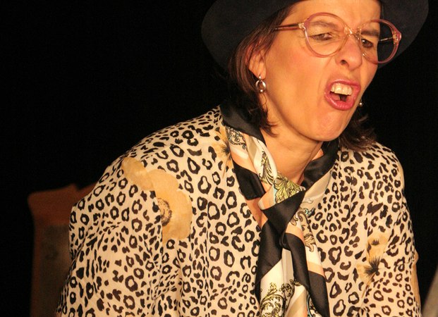 Eine Frau mit Brille und Hut mit gelber Blume zieht eine Grimasse. Sie trägt ein Leopardenmuster-Oberteil und einen Schal., © Galli Theater Backnang Eine Frau mit Brille und Hut mit gelber Blume zieht eine Grimasse. Sie trägt ein Leopardenmuster-Oberteil und einen Schal., © Galli Theater Backnang