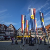 Der Marktplatz von Bad Urach zeigt Fachwerkhäuser und Fahnen. Menschen stehen auf dem gepflasterten Platz unter blauem Himmel., © Bad Urach Tourismus