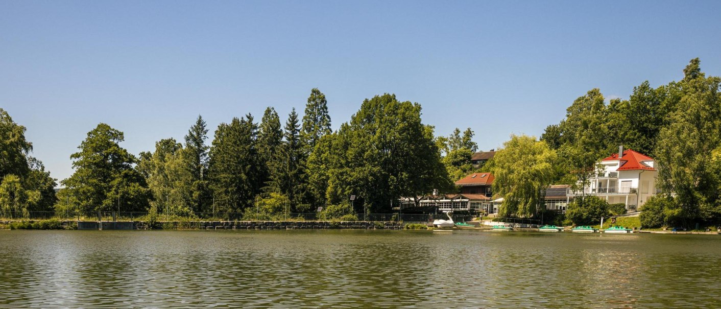Der Waldsee in Fornsbach mit umgebenden Bäumen und Gebäuden am Ufer unter klarem Himmel., © SMG, Sarah Schmid Der Waldsee in Fornsbach mit umgebenden Bäumen und Gebäuden am Ufer unter klarem Himmel., © SMG, Sarah Schmid