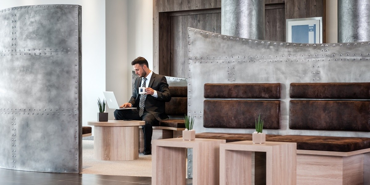 Ein Mann im Anzug arbeitet in einer modernen Lobby. Er hält eine Kaffeetasse und nutzt einen Laptop. Die Einrichtung ist stilvoll und minimalistisch., © Essential by Dorint