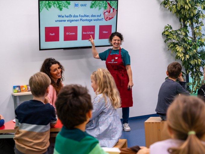 Eine Frau in roter Sch&uuml;rze pr&auml;sentiert ein Quiz auf einem Bildschirm vor einer Gruppe von Kindern in einem Klassenzimmer., &copy; Alfred Ritter GmbH &amp; Co. KG | Waldenbuch