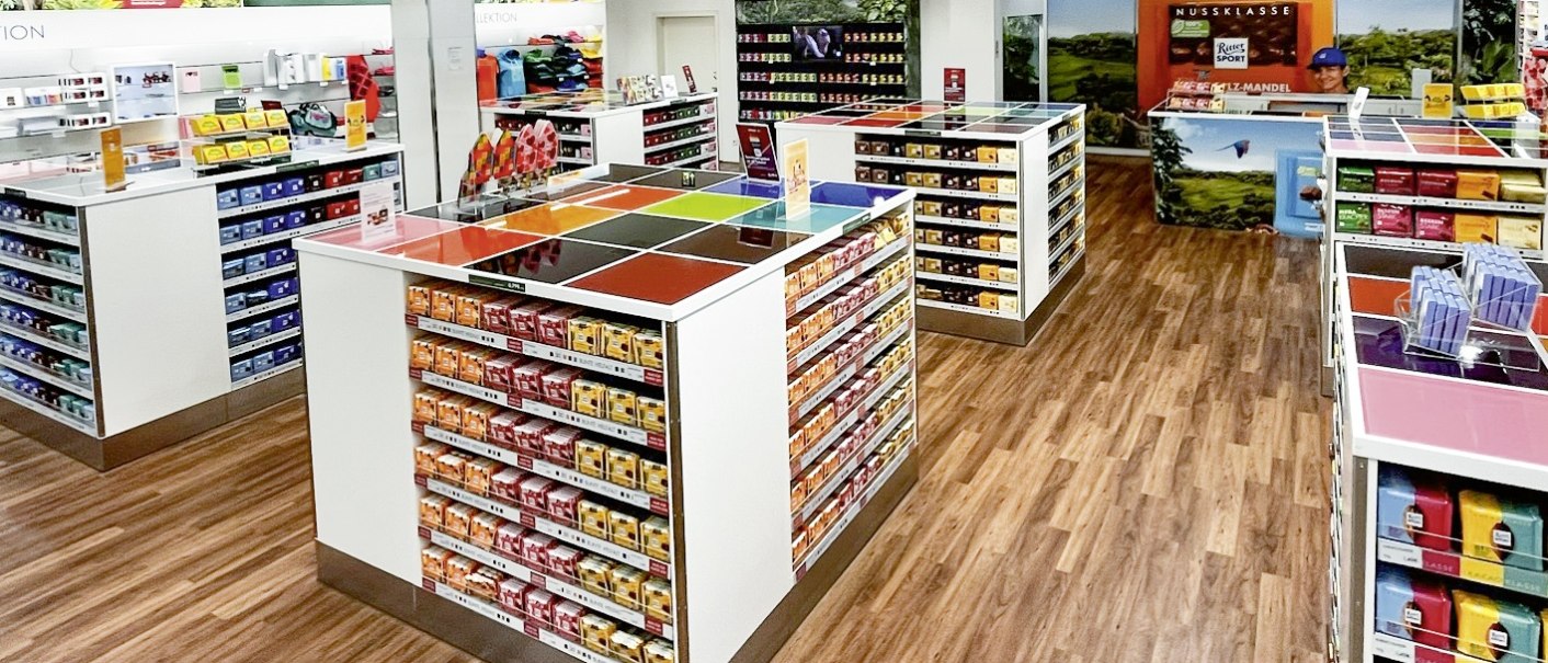 Ein heller Schokoshop mit bunten Ritter Sport Tafeln in Regalen, Holzboden und Werbepostern an den Wänden., © Alfred Ritter GmbH & Co. KG