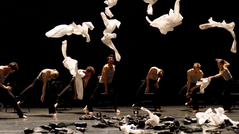 Tänzer auf einer dunklen Bühne werfen weiße Tücher in die Luft. Sie tragen schwarze Kleidung und bewegen sich synchron., © Regina Brocke Tänzer auf einer dunklen Bühne werfen weiße Tücher in die Luft. Sie tragen schwarze Kleidung und bewegen sich synchron., © Regina Brocke